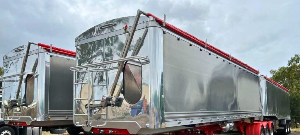 Polished aluminium bulk haulage trailers fitted with red rollover tarp systems parked outdoors under cloudy skies.